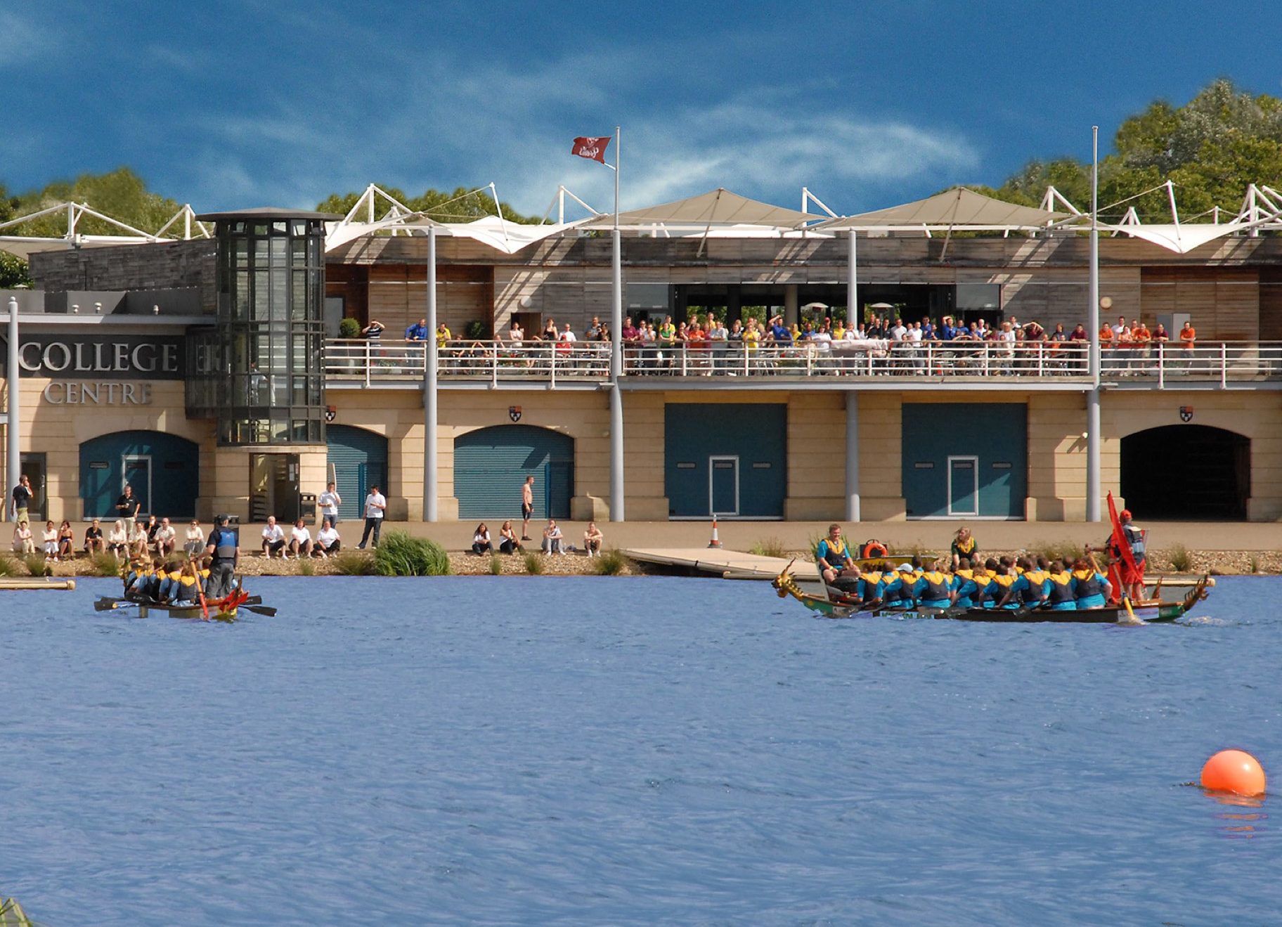 Corporate dragon boat racing event at waterside venue with teams competing and spectators watching from terrace
